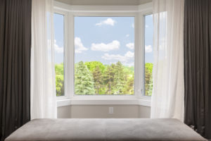A bay window flanked by dark grey and white drapes, with a view of a grove of trees outside